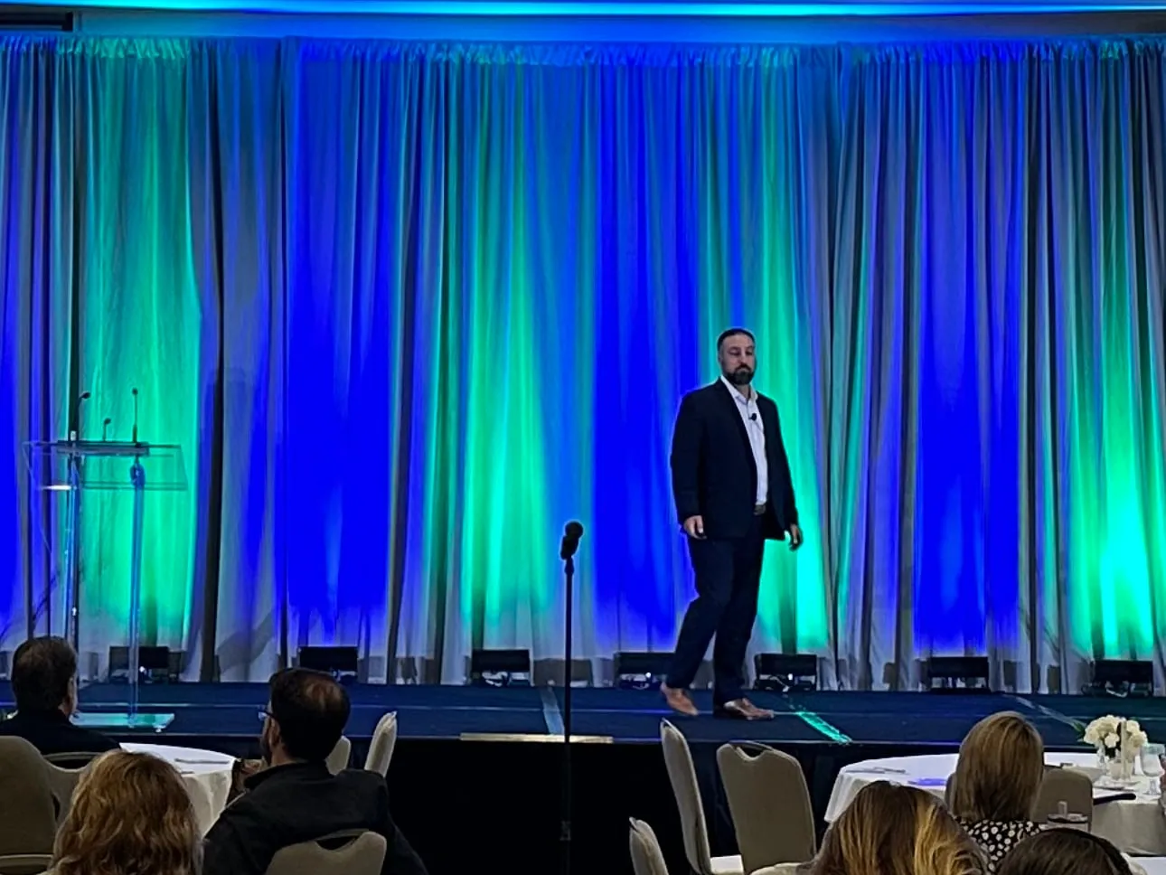 Hakim on a stage lit blue and green, with attendees at round tables watching from the front of the room