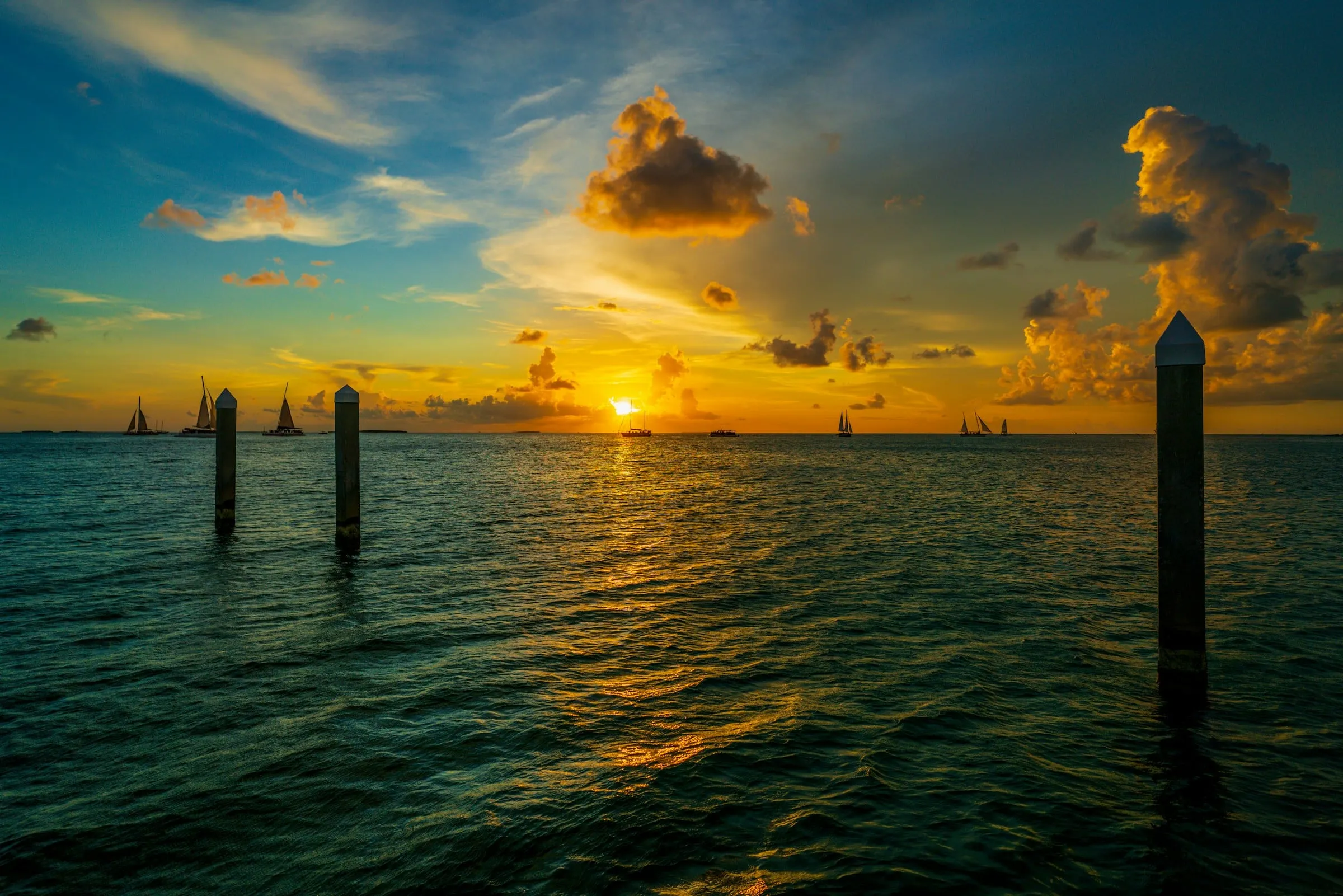 The sun setting over calm water in the Florida Keys, with sailboats on the horizon and dock pilings in the foreground